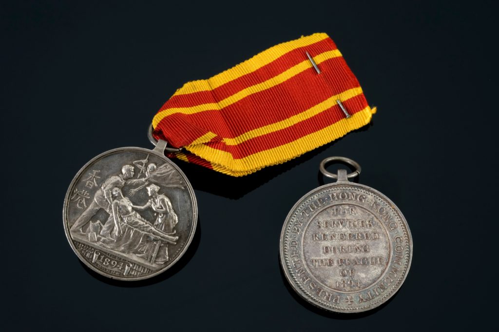 Two medals lying on a dark surface. One of them has a yellow and red ribbon attached, and depicts a man carrying another man on a stretcher and holding away the angel of death. A nurse leans towards the man on the stretcher. The year 1894 is displayed below. The other medal has the text “For services rendered during the plague of 1894’ surrounded by the text “Presented by the Hong Kong community” going around the edge.