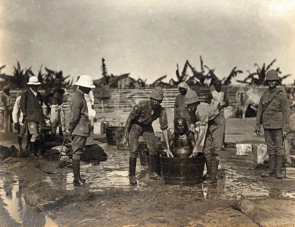 Monochrome photograph showing a group of British officers washing an Indian man in a wooden tub.