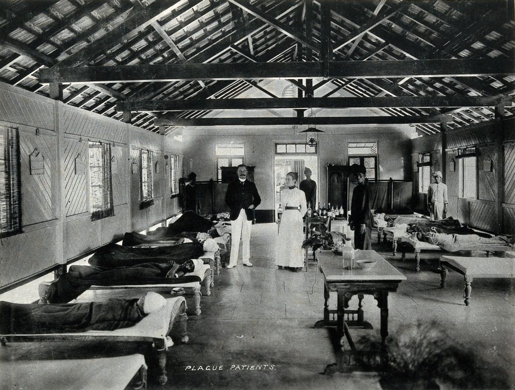 Monochrome photograph of a room lined with beds where people lie wrapped in blankets. A welldressed man and woman stand in the centre of the room facing the camera.